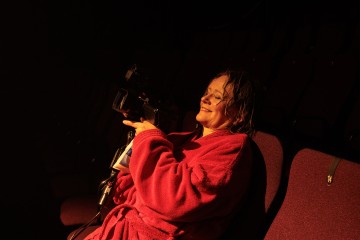 Punaiseen kaapuun pukeutunut nainen istuu hämärässä teatterissa hymyillen ja videokamera kädessään, ja hänen ympärillään on tyhjiä punaisia istuimia.