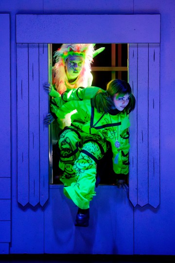 Kaksi ihmistä neonvihreissä puvuissa ja kasvomaalissa kiipeää lavalla olevan sarjakuvamaisen ikkunakehyksen läpi, jota valaisee mustavalo, joka antaa heidän asuilleen ja meikilleen hehkuvan vaikutelman.
