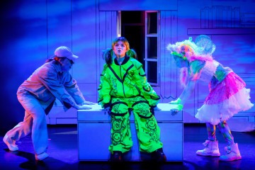 Lavalla esiintyy kolme ihmistä: yksi tavallisessa lippiksessä ja takissa, yksi neonvihreässä asussa, jossa on sarjakuvapiirroksia, ja yksi värikkäässä keijupuvussa. He ovat kokoontuneet valkoisen laatikon ympärille, jossa on sinisellä valaistu sarjakuvatausta.