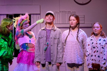 Viisi lasta seisoo lavalla rivissä, jotkut silmät kiinni. Yhdellä on värikäs, karvainen puku ja hän tökkii leikkisästi lippalakkipäisen pojan poskea. Muilla on yllään pyjama tai rennot vaatteet. Taustalla on yksinkertainen piirros huoneesta.