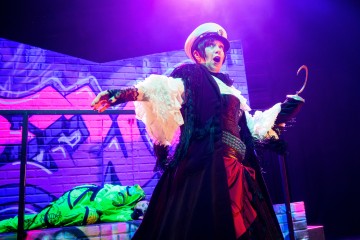 Dramaattiseen pukuun pukeutunut esiintyjä, jolla on valkoinen hattu ja koukkukäsi, seisoo lavalla yllättynyt ilme kasvoillaan. Taustalla näkyy värikkäitä graffiteja ja vihreään asuun pukeutunut henkilö violetissa valaistuksessa.