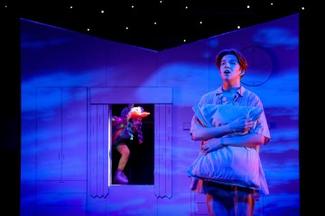 Pyjama-asuinen henkilö puristaa tyynyä ja katsoo ylöspäin tunnepitoinen ilme makuuhuoneen näköisellä lavasteella. Toinen värikkäästi pukeutunut esiintyjä kiipeää taustalla olevasta ikkunasta. Sininen valaistus luo unenomaista tunnelmaa.