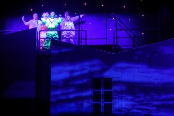 Neljä värikkäisiin pukuihin pukeutunutta näyttelijää esiintyy korotetulla näyttämöllä sinisessä ja violetissa valaistuksessa, ja lavasteisiin heijastetaan taustalle tähtiä ja pilviä.