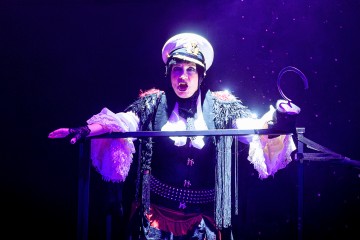 Dramaattiseen merirosvo-aiheiseen pukuun pukeutunut esiintyjä seisoo violettien näyttämövalojen alla, hänellä on valkoinen kapteeninhattu ja koukku kädessään, kädet ojennettuina ja ilmeikäs ilme kasvoillaan.