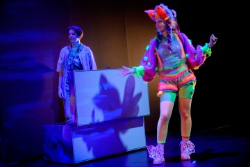 Lavalla on kaksi esiintyjää värikkään valaistuksen alla. Toinen seisoo hymyillen avoimen laatikon sisällä, kun taas toisella on kirkas, karvainen, värikäs puku, jossa on suuria rusetteja, ja hän poseeraa itsevarmasti edessä.