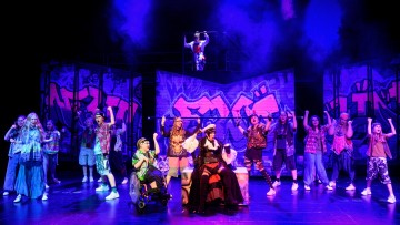 Monipuolinen joukko värikkäisiin pukuihin pukeutuneita esiintyjiä, joista yksi on pyörätuolissa ja yksi merirosvona, tanssii tarmokkaasti lavalla, jossa on graffiti-tyylisiä lavasteita ja dramaattinen violetti valaistus.