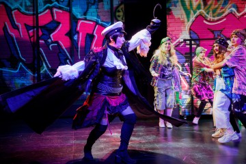 Dramaattiseen merirosvopukuun pukeutunut esiintyjä, jolla on koukkukäsi, kohtaa lavalla neljän henkilön ryhmän, joka on pukeutunut värikkäisiin, graffitista inspiroituneisiin asuihin, ja taustalla on kirkas graffitiseinä.