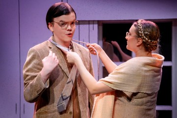 Nainen säätää miehen solmiota, kun tämä näyttää yllättyneeltä. Molemmilla on silmälasit ja vintage-tyyliset vaatteet, ja he seisovat kirkkaasti valaistussa huoneessa, jonka seinät ovat vaalean violetit ja jonka taustalla on ikkuna.