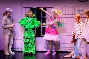 Neljä värikkäisiin pukuihin pukeutunutta ihmistä seisoo huonetta muistuttavalla lavasteella, ja yksi kirkkaanvihreä ja toinen vaaleanpunainen keijupukuinen henkilö elehtii innoissaan, kun taas kaksi muuta pyjama-asuisia katselee.