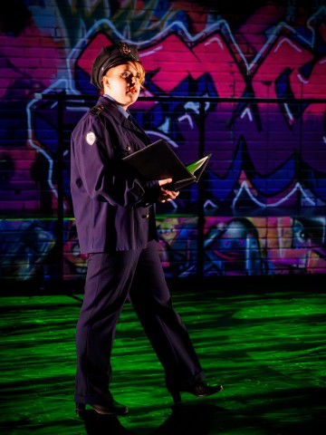 Poliisin univormuun pukeutunut henkilö seisoo pitelemässä avointa kansiota, värikäs graffititaide peittää hänen takanaan olevan tiiliseinän ja dramaattinen valaistus heittää varjoja lattialle.