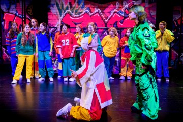 Ryhmä värikkäisiin pukuihin pukeutuneita nuoria esiintyjiä seisoo lavalla, jonka taustalla on graffitilla päällystetty tiiliseinä, kun taas kaksi kirkkaisiin asuihin pukeutunutta näyttelijää on dramaattisessa vuorovaikutuksessa keskellä.