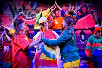 Värikkäisiin asuihin pukeutunut vilkas joukko nuoria antaa toisilleen lavalla korkeita vippauksia, ja taustalla on elävä graffitiseinä, joka luo juhlavan ja energisen ilmapiirin.