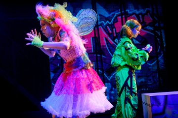 Lavalla esiintyy kaksi näyttelijää, joista toisella on värikkäät keijusiivet, vaaleanpunainen tutu ja karvainen liivi, kun taas toisella on kirkkaan vihreä asu. He seisovat eloisan graffititaiteen taustaa vasten.