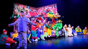 Joukko värikkäisiin pukuihin pukeutuneita lapsia istuu ja seisoo graffitilla päällystetyn tiiliseinän muotoisen lavastuksen ympärillä, kun taas harmaapukuinen aikuinen seisoo etukeskellä kädet ojennettuina kirkkaasti valaistulla näyttämöllä.