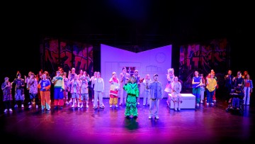 Suuri joukko esiintyjiä värikkäissä, eklektisissä puvuissa seisoo lavalla, jonka taustalla on värikkäitä graffiti-tyylisiä taustoja ja keskellä moderni, valkoinen lavaste. Kohtaus on kirkkaasti valaistu, mikä viittaa vilkkaaseen teatterituotantoon.