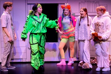 Lavalla seisoo viisi nuorta värikkäissä puvuissa. Yhdellä on kirkkaanvihreä asu, ja hän osoittaa eleillään muille, jotka reagoivat yllättyneinä ja innostuneina. Tausta viittaa teatterinäytelmään, jossa on yksinkertainen lavastus.