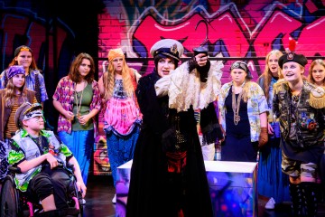 Kapteeni Koukuksi pukeutunut henkilö esiintyy dramaattisesti lavalla värikkäisiin pukuihin pukeutuneiden lasten, joista yksi on pyörätuolissa, ympäröimänä.