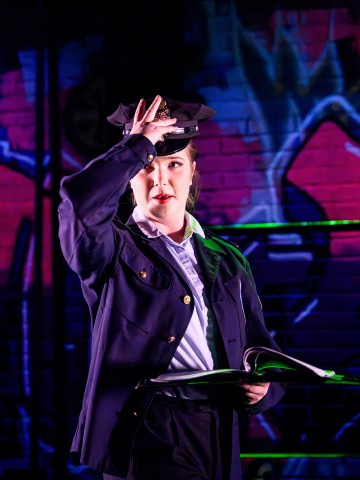 Poliisin univormuun pukeutunut henkilö pitää kirjaa kädessään ja säätää hattua seisoen kirkkaanvärisen, graffitilla päällystetyn seinän edessä.