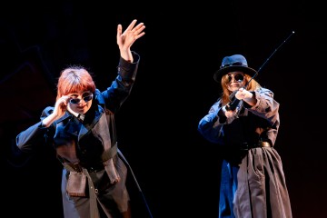 Lavalla esiintyy kaksi ihmistä, joilla on trenssitakit, hatut ja aurinkolasit. Toinen nostaa kättä dramaattisesti, kun taas toinen osoittaa kapulaa eteenpäin. Molemmat seisovat kirkkaassa valaistuksessa tummaa taustaa vasten.