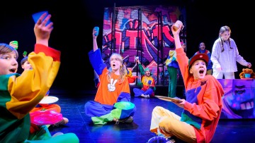 Värikkäisiin asuihin pukeutuneet lapset istuvat lavalla ja nostelevat innoissaan kuppeja ja lautasia. Taustalla pöydän ääressä seisoo tyttö. Heidän takanaan on kirkkaasti maalattu graffitityylinen tausta. Kohtaus on vilkas ja teatraalinen.