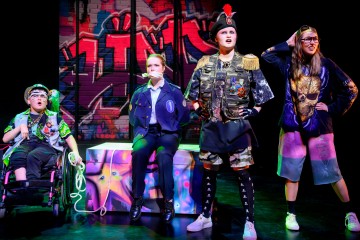 Neljä näyttelijää esiintyy lavalla graffititaiteen edessä. Yksi istuu pyörätuolissa köysi kädessä, yksi on sidottu tuoliin poliisin univormussa, yhdellä on armeijan asu ja yksi pukeutuu rentoihin vaatteisiin, joissa on leijonagrafiikka.