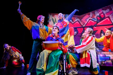 Lavalla esiintyy värikkäisiin pukuihin pukeutunut vilkas joukko näyttelijöitä; yksi pyörätuolissa istuva henkilö pitelee suurta sekoitusastiaa, ja muut poseeraavat iloisesti heidän ympärillään eloisan graffitityylisen taustan edessä.