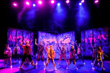 Ryhmä esiintyjiä tanssii lavalla värikkäiden violettien ja keltaisten valojen alla, ja taustalla on värikkäitä graffititauluja. Esiintyjillä on rennot, urbaanin tyyliset puvut, ja he poseeraavat energisesti laajassa muodostelmassa.