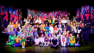 Suuri joukko värikkäisiin pukuihin ja rentoihin vaatteisiin pukeutuneita ihmisiä poseeraa yhdessä näyttämöllä, jonka taustalla on elävä, graffitilla peitetty tausta, hymyillen ja juhlien sitä, mikä näyttää olevan teatterituotanto.