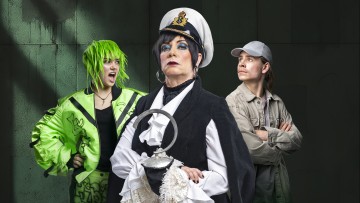 Kolme ihmistä poseeraa dramaattisesti: kapteeninhattuun ja mustaan viittaan pukeutunut vakava vanhempi henkilö pitelee hopeista koukkua, vasemmalla kirkkaanvihreä tukkainen ja neonväriseen takkiin pukeutunut nuorempi henkilö huutaa ja oikealla toinen seisoo kädet ristissä.