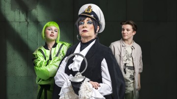 Kolme ihmistä poseeraa puku päällä tummaa, kuvioitua seinää vasten. Keskushenkilöllä on merisotilashattu ja hänellä on koukku kädessään, ja häntä reunustavat vasemmalla neonvihreään peruukkiin ja asuun pukeutunut henkilö ja oikealla rentoon beigeen pukeutunut henkilö.