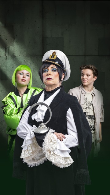 Kolme ihmistä poseeraa puvuissa: edessä olevalla henkilöllä on dramaattinen meikki, valkoinen merivoimien hattu ja mustavalkoinen asu; heidän takanaan toisella henkilöllä on neonvihreä tukka ja kirkas takki, toisella on lyhyet hiukset ja rento vaatetus.