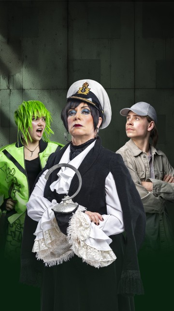 Kolme ihmistä poseeraa tummaa taustaa vasten: edessä seisoo kapteeninhattuun ja dramaattiseen meikkiin pukeutunut nainen, jota reunustaa neonvihreä tukka ja takki sekä sivuttain katsova harmaaseen hattuun ja takkiin pukeutunut henkilö.