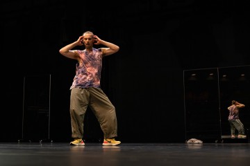 Henkilö, jolla on hihaton paita ja löysät housut, seisoo lavalla kädet päänsä päällä. Taustalla näkyy hänen peilikuvansa peilistä. Lava on hämärästi valaistu, ja lattialla on hattu tai kangas.