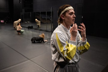 Verryttelypukuun ja otsapantaan pukeutunut henkilö seisoo keskittyneenä etualalla tummalla näyttämöllä, kun taas kolme muuta henkilöä tekee ilmeikkäitä, vaihtelevia poseerauksia taustalla, osa lattialla, ja heidän takanaan on peiliseinä.