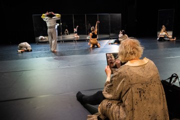 Henkilö istuu lattialla meikkaamassa ja katsoo peiliin, kun useat tanssijat esiintyvät eri asennoissa suurten peilien edessä hämärästi valaistulla näyttämöllä.