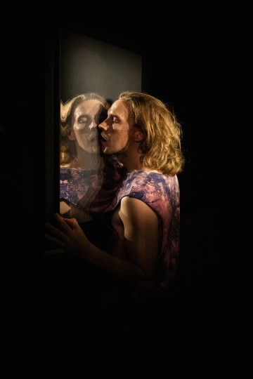 Henkilö, jolla on pitkät vaaleat hiukset ja kuvioitu hihaton paita, kumartuu suutelemaan omaa peilikuvaansa pimeässä peilissä, jota valaisee dramaattinen valaistus.