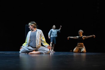 Kolme esiintyjää poseeraa tummalla näyttämöllä ilmeikkäissä asennoissa; yksi istuu ristissä etualalla, toinen seisoo paljain jaloin takana ja kolmas polvistuu kädet kohotettuna luoden dramaattisen kohtauksen.