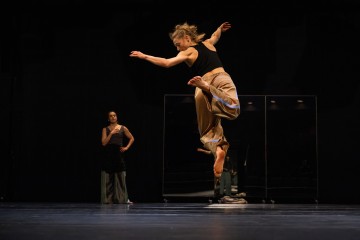 Paljasjalkainen tanssija, jolla on ruskehtavat housut ja musta toppi, hyppii sulavasti ilmassa hämärästi valaistulla näyttämöllä, kun toinen henkilö seisoo taustalla tarkkailemassa.