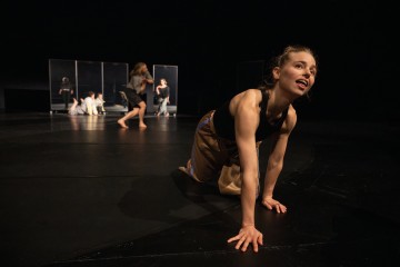 Ruskeaan mekkoon pukeutunut nainen polvistuu tummalla näyttämöllä, nojaa eteenpäin, toinen käsi lattialla, ja katsoo ylöspäin. Taustalla on useita muita tanssijoita peilipaneelien lähellä.