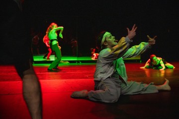 Harmaisiin vaatteisiin pukeutunut henkilö polvistuu hämärästi valaistulla näyttämöllä ja elehtii ilmeikkäästi käsillään. Hänen takanaan liikkuu muita dynaamisissa asennoissa olevia esiintyjiä dramaattisessa punaisessa ja vihreässä valaistuksessa.