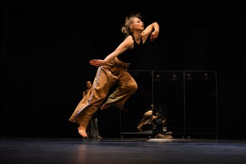 Tanssija, jolla on löysät ruskeat housut ja musta toppi, hyppää sirosti lavalla, toinen käsi ylöspäin taivutettuna, tummaa taustaa vasten, jossa peili heijastaa hänen liikkeensä.