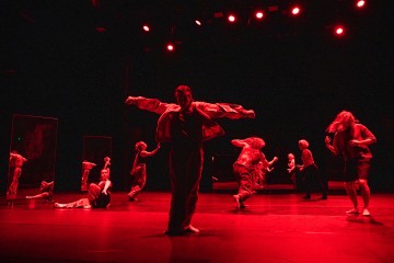 Ryhmä tanssijoita esiintyy lavalla dramaattisessa punaisessa valaistuksessa. Jotkut tanssijat seisovat kädet ojennettuina, toiset istuvat tai liikkuvat energisesti luoden dynaamisen, ilmeikkään kohtauksen. Taustalla näkyy korkeita peilejä.