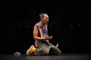 Henkilö, jolla on pitkät hiukset pullana, hihaton solmio-paita, lasti-housut ja keltaiset kengät, polvistuu tummalla näyttämöllä, elehtii yhdellä kädellä ja näyttää olevan syvällä ajatuksissaan tai tunteissaan.