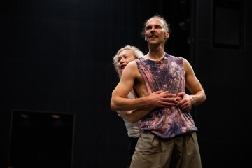 Kaksi ihmistä seisoo lähekkäin lavalla; valkopukuinen nainen halaa miestä takaapäin. Mies, jolla on yllään hihaton violetti paita ja khakihousut, katsoo hieman hymyillen eteenpäin. Tausta on tumma, mikä viittaa teatteriympäristöön.