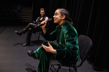 Vihreään samettipukuun pukeutunut henkilö, jolla on punotut hiukset, puhuu mikrofoniin lavalla istuen. Toinen mustiin pukeutunut henkilö istuu kauempana hymyillen tummien verhojen taustaa vasten.