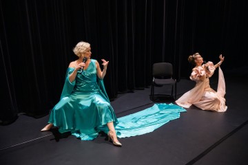 Lavalla istuu kaksi tyylikkääseen pukuun pukeutunutta naista mikrofonien kanssa, toinen teerenvärinen tuolilla, jossa on pitkä juna, ja toinen vaaleanpunainen lattialla, ja he elehtivät dramaattisesti mustien verhojen taustaa vasten.