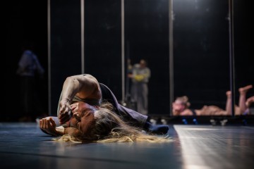 Henkilö makaa kyljellään hämärästi valaistulla näyttämöllä, toinen käsi taivutettuna kasvojensa lähelle ja intensiivinen ilme kasvoillaan. Taustalla seisoo kaksi ihmistä, ja toinen henkilö makaa, kaikki hieman epätarkasti.