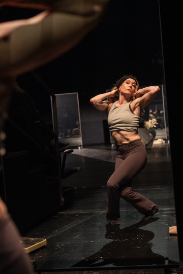 Urheiluvaatteisiin pukeutunut nainen poseeraa lavalla itsevarmasti, toinen polvi koukussa ja kädet pään taakse nostettuina, ja hänen kätensä heijastuvat läheisestä peilistä. Kohtaus on hämärästi valaistu, ja taustalla näkyy näyttämölaitteita.