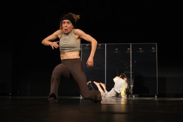 Harmaaseen crop-toppiin ja ruskeisiin housuihin pukeutunut nainen polvistuu lavalla yllättynyt ilme kasvoillaan, kun taas kaksi muuta vaaleisiin vaatteisiin pukeutunutta ihmistä on sumeana taustalla läpinäkyvän väliseinän lähellä.