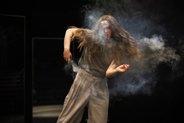 Lavalla tanssii pitkähiuksinen ja löysästi pukeutunut henkilö, jota ympäröi valkoinen savupilvi ja jonka tausta on tumma.